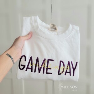 Pu&ograve; includere: Felpa bianca con le parole "GAME DAY" ricamate in viola e "Vikings" in oro. Il logo Wild Son Goods &egrave; visibile in basso a destra.