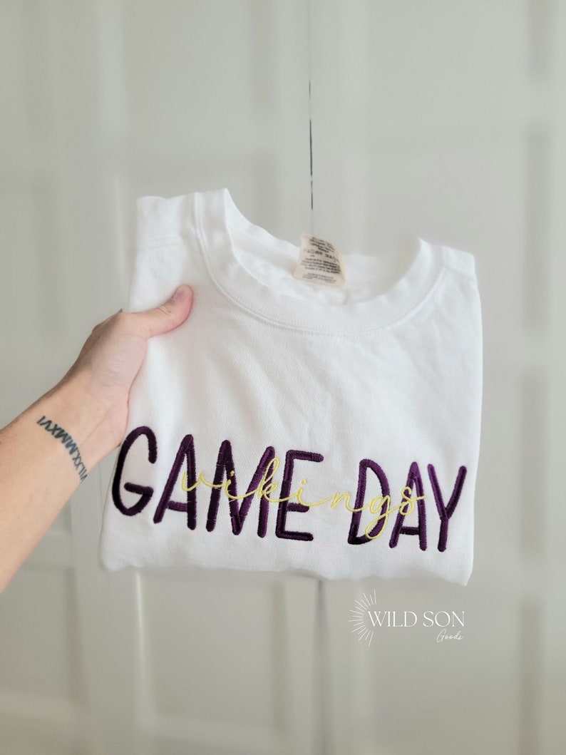 Pu&ograve; includere: Felpa bianca con le parole "GAME DAY" ricamate in viola e "Vikings" in oro. Il logo Wild Son Goods &egrave; visibile in basso a destra.