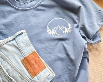 May include: A light blue t-shirt with a white mountain and tree design, paired with light wash denim shorts. The shirt has a Comfort Colors tag. A brown leather patch is visible on the shorts.