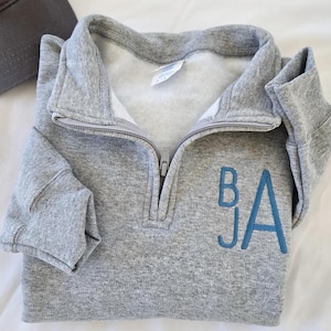 Könnte beinhalten: Ein graues Sweatshirt mit Viertelreißverschluss und den gestickten Initialen "BJA" in Blau. Das Sweatshirt hat einen gefalteten Kragen und eine teilweise geöffneten Reißverschluss. Eine graue Baseballkappe ist oben links zu sehen.