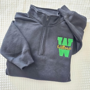 May include: A black quarter-zip sweatshirt with a green and yellow embroidered patch that reads "Willow" with a "W" design. The sweatshirt has a zipper closure and a hood.