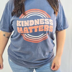 Puede incluir: Una camiseta azul con las palabras "KINDNESS MATTERS" en blanco y naranja, enmarcada por un dise&ntilde;o de arco&iacute;ris retro. La camiseta de manga corta se combina con vaqueros azules, creando un look casual.