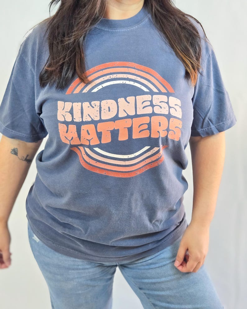 Puede incluir: Una camiseta azul con las palabras "KINDNESS MATTERS" en blanco y naranja, enmarcada por un dise&ntilde;o de arco&iacute;ris retro. La camiseta de manga corta se combina con vaqueros azules, creando un look casual.