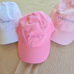 May include: Three baseball caps in white and pink. The white cap has the name "Beau" embroidered in blue. The pink caps have the names "Jocelyn" and "Margaret" embroidered in blue. All caps have a curved brim and a button on top.