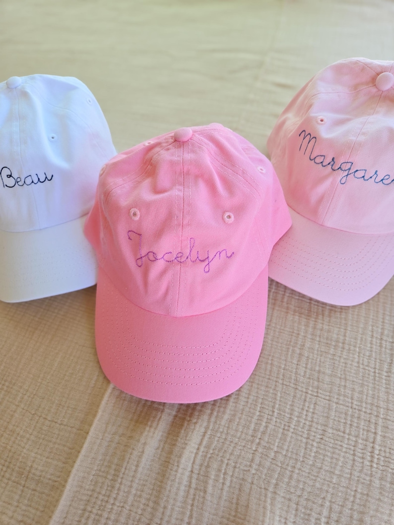 May include: Three baseball caps in white and pink. The white cap has the name "Beau" embroidered in blue. The pink caps have the names "Jocelyn" and "Margaret" embroidered in blue. All caps have a curved brim and a button on top.