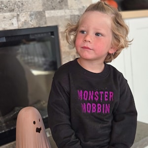 May include: A child wearing a black sweatshirt with the text "MONSTER MOBBIN" in purple lettering. The child is wearing orange pants and is next to a peach-colored ghost decoration. The background includes a fireplace and a stone wall.