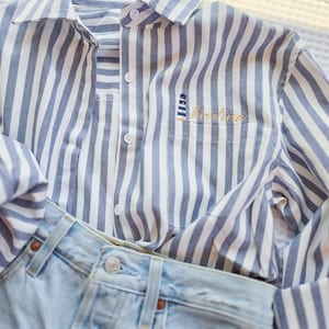 Może przedstawiać: Niebiesko-biała koszula w paski z kieszenią z haftowaną latarnią morską i imieniem "Kristina" złotą nicią. Widoczne są jasnoniebieskie dżinsy. Koszula ma długie rękawy.