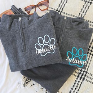 Peut inclure: Deux sweat-shirts gris à fermeture éclair quart de zip avec des empreintes de pattes et des noms brodés. L'un a une empreinte de patte bleu clair avec le nom "Danielle", et l'autre a une empreinte de patte turquoise avec le nom "Autumn".