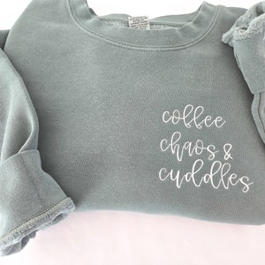 May include: Sage green sweatshirt with the words "coffee chaos & cuddles" in white script. The long-sleeved top has a crew neck and ribbed cuffs. The fabric appears soft and comfortable.