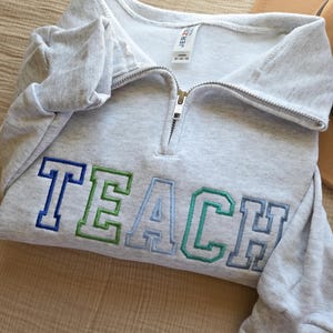 Puede incluir: Una sudadera gris claro con cuello con cremallera. La palabra "TEACH" est&aacute; bordada en la parte delantera en azul, verde y azul claro. La sudadera est&aacute; doblada y parece estar hecha de un material suave.