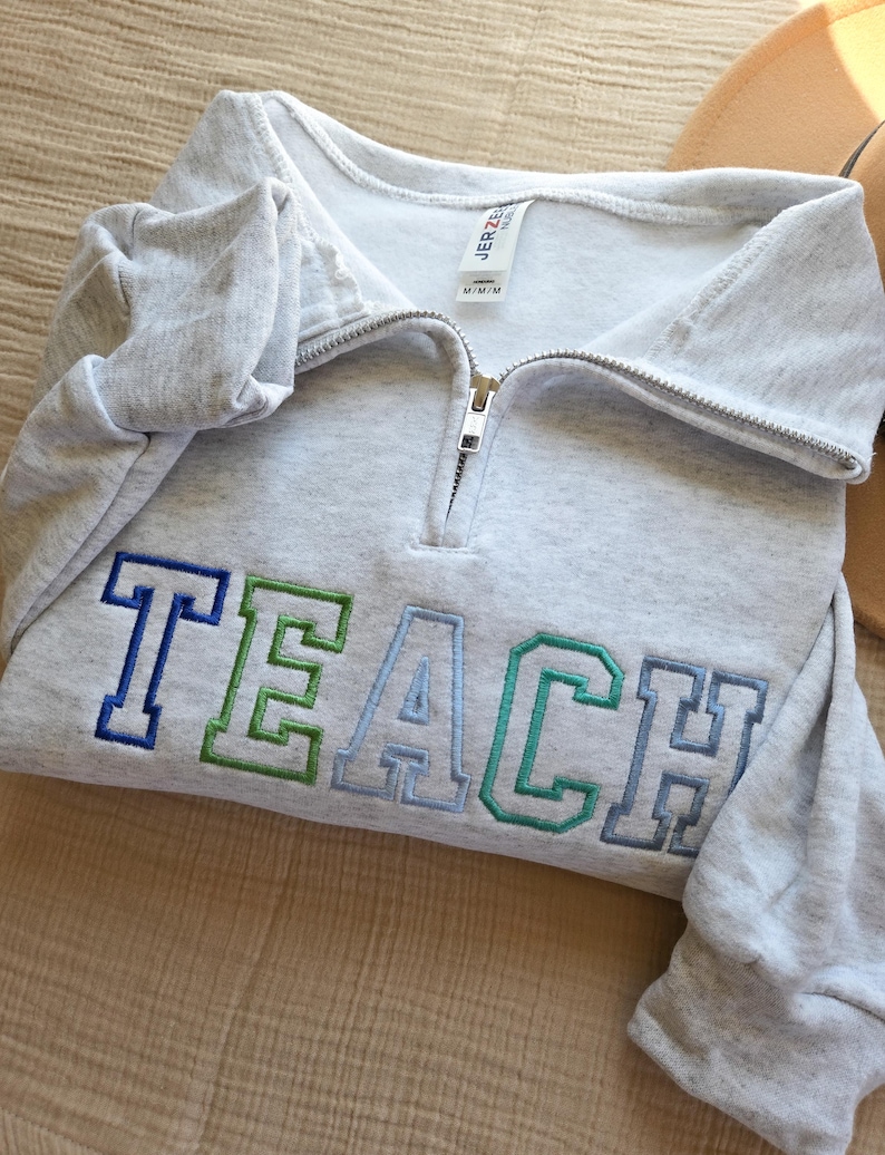 Puede incluir: Una sudadera gris claro con cuello con cremallera. La palabra "TEACH" est&aacute; bordada en la parte delantera en azul, verde y azul claro. La sudadera est&aacute; doblada y parece estar hecha de un material suave.