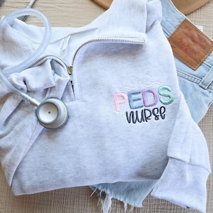 Könnte beinhalten: Ein hellgraues Sweatshirt mit Viertelreißverschluss und dem gestickten Text "PEDS nurse" in Pastellfarben. Ein Stethoskop liegt auf dem Sweatshirt, das mit Jeansshorts und einem braunen Hut kombiniert wird.
