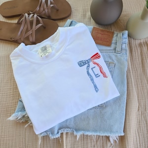 May include: A white t-shirt with an embroidered design in red, white, and blue, alongside light-wash denim shorts and brown sandals. The items are arranged on a neutral fabric, with small vases of flowers. The t-shirt is from Comfort Colors.