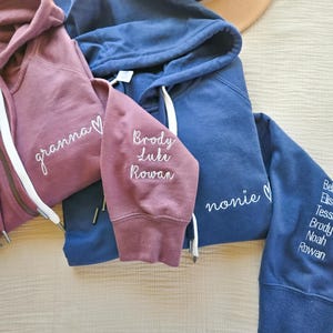 May include: Three hooded sweatshirts in shades of dusty rose and navy blue. The dusty rose sweatshirt has the word "granna" embroidered on the front. The navy blue sweatshirts have the words "nonie" and names embroidered on the sleeves.