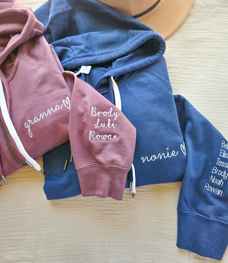 May include: Three hooded sweatshirts in shades of dusty rose and navy blue. The dusty rose sweatshirt has the word "granna" embroidered on the front. The navy blue sweatshirts have the words "nonie" and names embroidered on the sleeves.
