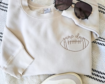 Può includere: Una felpa color crema con la scritta ricamata "game day" e una grafica di football. Occhiali da sole neri poggiano sulla felpa. Scarpe da ginnastica bianche con strisce nere nella parte inferiore dell'immagine.