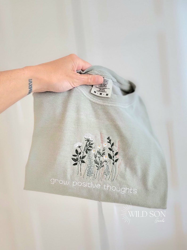 Puede incluir: Sudadera verde salvia con dise&ntilde;o floral bordado y la frase "grow positive thoughts". El bordado presenta flores y follaje blancos, rosas y verdes. La sudadera est&aacute; doblada.