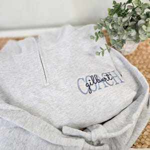 May include: A light gray heathered quarter-zip sweatshirt featuring embroidered text on the chest. The word COACH is stitched in large light blue block letters, with the name gilbert embroidered in black cursive script layered over it.