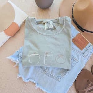 May include: A sage green t-shirt with embroidered kitchen-themed designs, including a pot, jar, and loaf of bread, is folded on top of light blue denim shorts. A beige hat, sandals, and a white and peach-colored clutch are also visible.