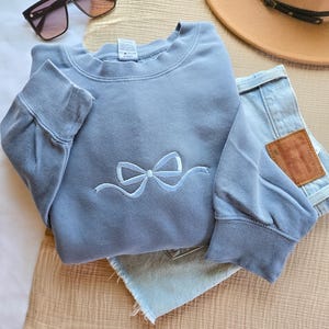 May include: A light blue sweatshirt with a bow design, paired with light wash denim shorts, sunglasses, and a brown hat. The sweatshirt features a crew neck and long sleeves. The bow is embroidered in a lighter shade of blue.