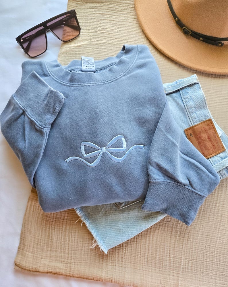 May include: A light blue sweatshirt with a bow design, paired with light wash denim shorts, sunglasses, and a brown hat. The sweatshirt features a crew neck and long sleeves. The bow is embroidered in a lighter shade of blue.