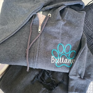 May include: A gray zip-up hoodie with a teal paw print and the name "Brittany" embroidered on the front. The hoodie has a drawstring hood and a zipper closure. Dark gray denim jeans are folded next to the hoodie.