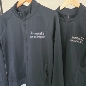 May include: Two black zip-up jackets hanging on a rail. Each jacket features the name "Jennifer" and the words "Dental Hygienist" printed in white, along with a tooth graphic. The jackets have pockets and a high collar.