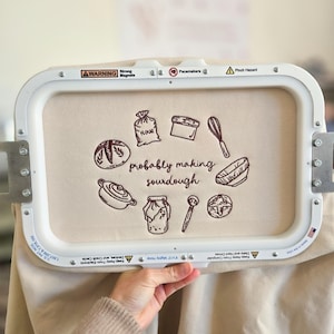 May include: An embroidery hoop with a beige fabric featuring brown illustrations of baking tools and bread, with the text "probably making sourdough." The design includes a flour sack, loaf of bread, and a whisk. The hoop is held by a hand.