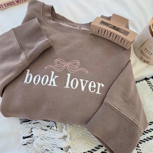 Op de afbeelding: Een bruine sweatshirt met de geborduurde tekst "book lover" en een roze strik. Een beige haarclip ligt op de sweatshirt. Een boek en een kaars zijn zichtbaar op de achtergrond.