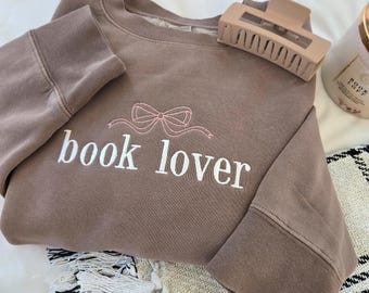 Könnte beinhalten: Ein braunes Sweatshirt mit dem gestickten Schriftzug "book lover" und einer rosa Schleife. Eine hellbraune Haarspange liegt auf dem Sweatshirt. Ein Buch und eine Kerze sind im Hintergrund zu sehen.