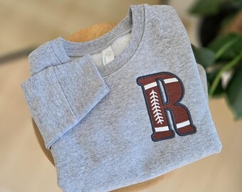 May include: A gray sweatshirt with a football-themed letter "R" patch in brown and white. The sweatshirt is folded and resting on a light-colored surface, with a blurred background.
