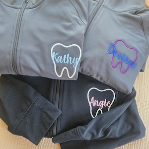 Könnte beinhalten: Drei Reißverschlussjacken in Grau und Schwarz. Jede Jacke hat ein gesticktes Zahndesign mit einem Namen: "Kathy" in Blau, "Desiree" in Blau und Lila und "Angie" in Rosa. Die Jacken sind aus weichem, sportlichem Material.