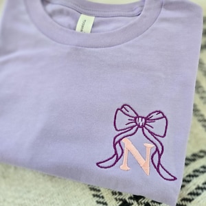Op de afbeelding: Een lila T-shirt met een ronde hals. Het shirt heeft een geborduurd strikdesign in donkerpaars met de letter "N" in lichtroze. Het shirt is opgevouwen en ligt op een zwart-wit patroon oppervlak.