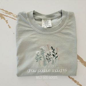 Puede incluir: Una camiseta verde salvia con cuello redondo y dise&ntilde;o floral bordado. La camiseta presenta el texto blanco "grow positive thoughts" y "WILD SON GOODS". La camiseta est&aacute; doblada cuidadosamente.