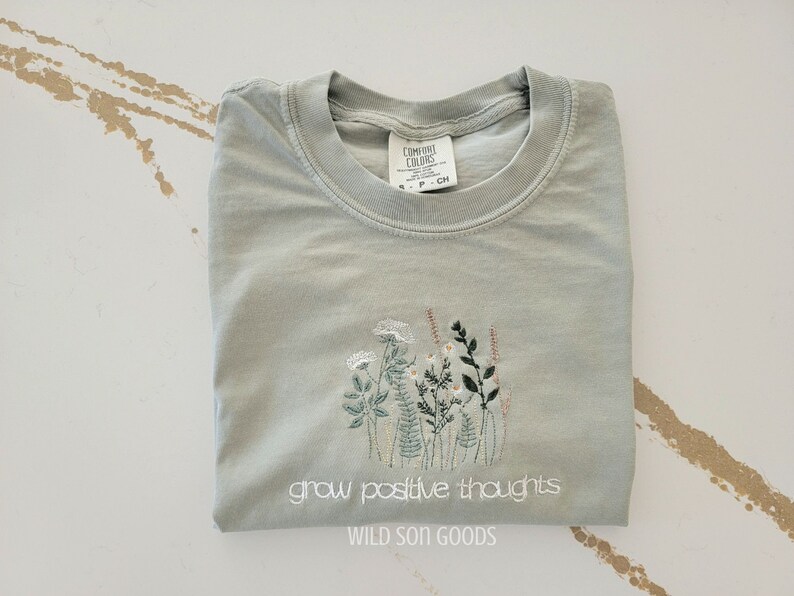 Puede incluir: Una camiseta verde salvia con cuello redondo y dise&ntilde;o floral bordado. La camiseta presenta el texto blanco "grow positive thoughts" y "WILD SON GOODS". La camiseta est&aacute; doblada cuidadosamente.
