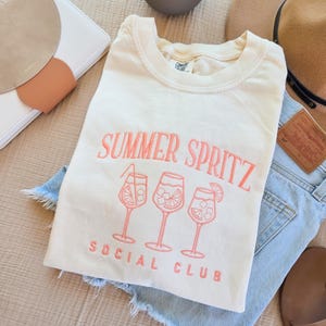 May include: Cream-colored t-shirt with the words "SUMMER SPRITZ SOCIAL CLUB" embroidered in coral. The design includes three cocktail glasses. The shirt is laid on top of denim shorts, with a hat and other accessories in the background.