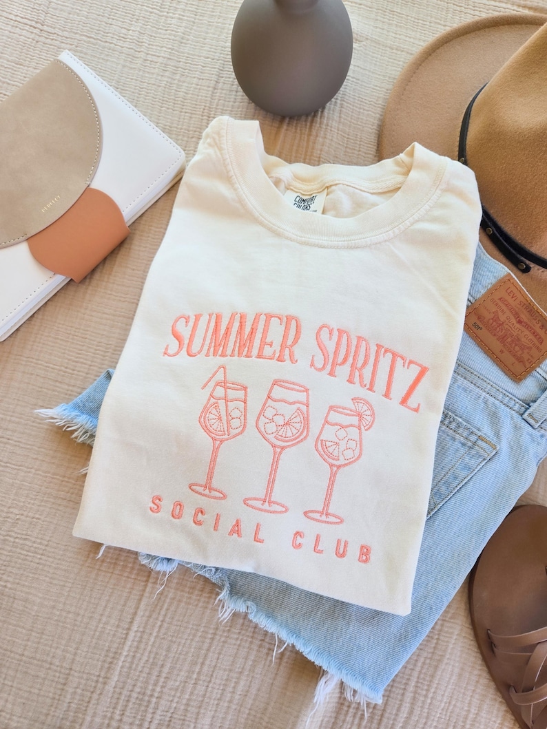 May include: Cream-colored t-shirt with the words "SUMMER SPRITZ SOCIAL CLUB" embroidered in coral. The design includes three cocktail glasses. The shirt is laid on top of denim shorts, with a hat and other accessories in the background.