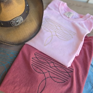 Puede incluir: Un sombrero de vaquero de paja con una banda de cuero marrón y hebilla plateada descansa junto a tres camisetas. Una camiseta rosa tiene un diseño bordado dorado, y una camiseta granate tiene un diseño bordado negro. También se ven unos vaqueros azules.