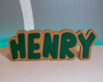 Könnte beinhalten: Ein hölzernes Namensschild mit der Aufschrift "HENRY" in Grün und Braun.