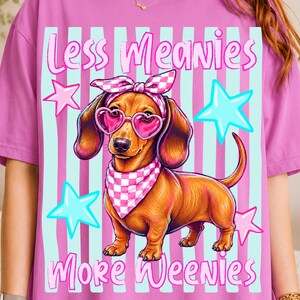 Könnte beinhalten: Ein pinkfarbenes T-Shirt mit dem Text "Less Meanies More Weenies" und einem Cartoon-Dackel mit herzförmiger Sonnenbrille, Bandana und Schleife. Das Design umfasst blaue Sterne und vertikale Streifen.