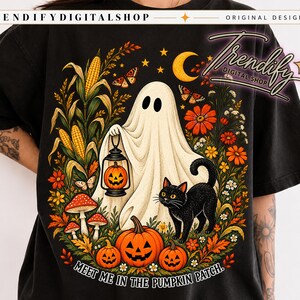 May include: A black t-shirt featuring a graphic of a white ghost holding a lantern with a jack-o'-lantern inside, a black cat, pumpkins, corn stalks, mushrooms, and orange flowers. The text "MEET ME IN THE PUMPKIN PATCH" is printed at the bottom.