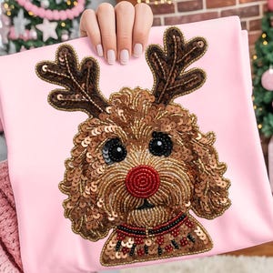 Peut inclure: Un coussin rose clair avec un motif de chien aux bois de renne en sequins et perles. Le chien a un nez rouge, des yeux noirs et un pelage brun. Le coussin est tenu par une personne.