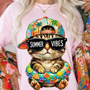 May include: Pink t-shirt featuring a cartoon cat wearing sunglasses with the words "Summer Vibes." The cat is wearing a colorful hat and a floatie. The design is vibrant and playful, perfect for summer.