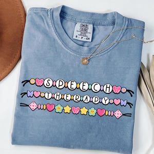 May include: A blue Comfort Colors t-shirt with the words "SPEECH THERAPY" in a beaded design. The design includes hearts, stars, and bows. A gold necklace with a pendant is draped across the shirt.