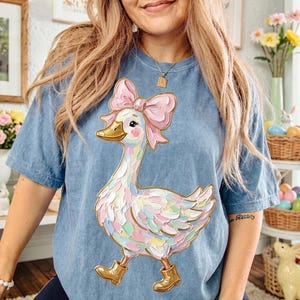 May include: A light blue t-shirt featuring a whimsical goose illustration. The goose has a pink bow, gold boots, and colorful feathers. The shirt is a comfortable, casual style, perfect for everyday wear.