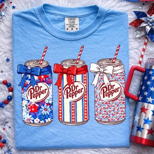 May include: Light blue Comfort Colors t-shirt featuring three Dr. Pepper soda can designs. Each can has a different patriotic floral or striped pattern, a bow, and a red and white striped straw. The shirt is a comfortable, casual style.
