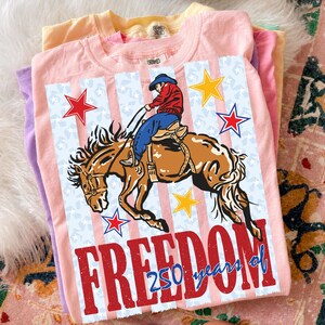 May include: A light pink t-shirt featuring a graphic of a cowboy riding a bucking horse. The design includes red, white, and blue vertical stripes, stars, and the text "FREEDOM 250 years of" in large red and blue lettering across the bottom.