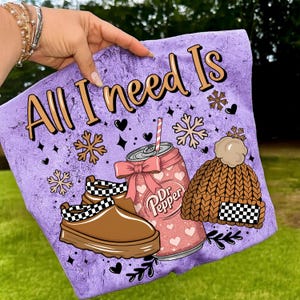 May include: A purple graphic featuring the text "All I need Is" with illustrations of a Dr. Pepper can, a brown knit hat, and brown shoes with a black and white checkered pattern. The design includes snowflakes and hearts.