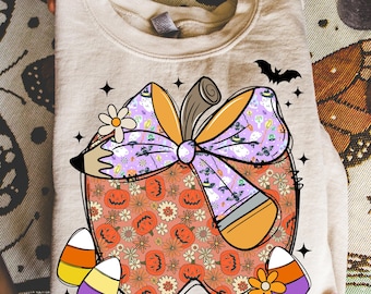 May include: Beige sweatshirt featuring a pumpkin design with a bow, pencil, and candy corn accents. The pumpkin has a floral pattern with jack-o'-lanterns. A bat and stars complete the Halloween-themed design.
