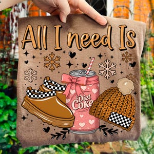 Könnte beinhalten: Braunes T-Shirt mit dem Text "All I need Is". Die Grafik zeigt eine rosa Diet Coke-Dose, braune Schuhe mit schwarz-weißem Schachbrettmuster und eine braune Strickmütze mit Schachbrettmuster.